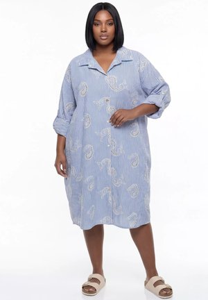 Kobieta w luźnej niebiesko-białej sukience w paski z wzorami paisley, stoi z jedną uniesioną ręką, beżowe sandały, neutralne tło.