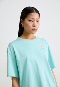 Tricou scurt cu mânecă scurtă, de culoare albastru deschis, fabricat din bumbac moale, având un grafic mic roz pe pieptul stâng și o croială lejeră.