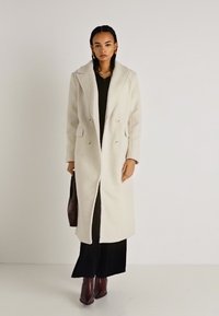 Dvojřadový, dlouhý krémový kabát s texturovanou tkaninou, klopovým límcem a bočními kapsami, nošený přes černý outfit a spárovaný s hnědými botami.