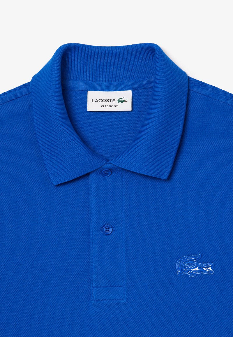 Blaues Poloshirt aus strukturiertem Baumwollmaterial, mit klassischem Schnitt. Beinhaltet eine Knopfleiste mit zwei Knöpfen und ein gesticktes Krokodil-Logo auf der Brust.