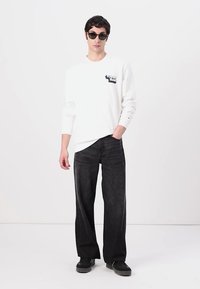 Hvid langærmet skjorte med et marineblåt logo, parret med brede sorte jeans og sorte sko. Beklædningen har en afslappet pasform og et enkelt design.