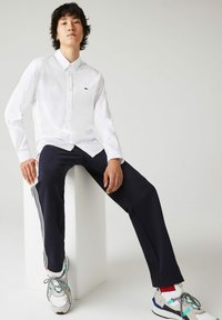 Witte lange mouwen button-up shirt, marineblauwe broek met witte strepen, en multicolor sneakers. Model zit op een witte pedestaal.