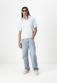 Ανοικτού μπλε textured polo shirt με κοντά μανίκια, συνδυασμένο με ελαφρώς φθαρμένα τζιν ίσιας γραμμής και μπεζ αθλητικά παπούτσια. Φορούνται μαύρα γυαλιά ηλίου.