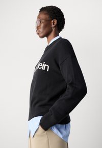 Frau mit kurzen, lockigen Haaren, die eine Brille trägt, einen schwarzen Pullover mit weißem Text, ein blaues Hemd und beige Hosen anhat, seitlich steht und die Hände in den Taschen hat.
