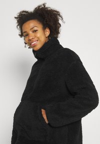 Pull en polaire noir avec col haut et manches longues. Présente une surface douce et texturée, ainsi que des poches latérales pour une fonctionnalité accrue.