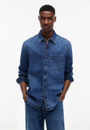 Młody mężczyzna z krótkimi włosami, ubrany w niebieską dżinsową koszulę na guziki z podwiniętymi rękawami oraz pasujące niebieskie dżinsy, z rękami w kieszeniach.