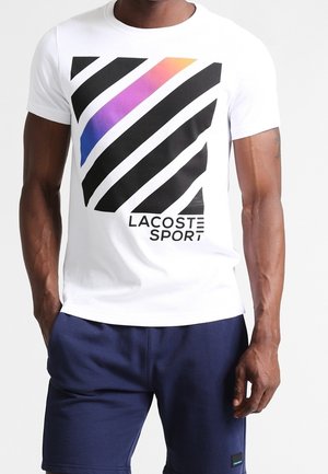 Homme portant un t-shirt blanc avec des rayures diagonales noires et un dégradé coloré, et un short bleu marine.