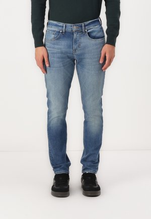 Modre jeans hlače z ozkim krojem, bledim pranjem in vidnimi šivi. Imajo pet žepov in zanke za pas, v kombinaciji z črnimi supergami.