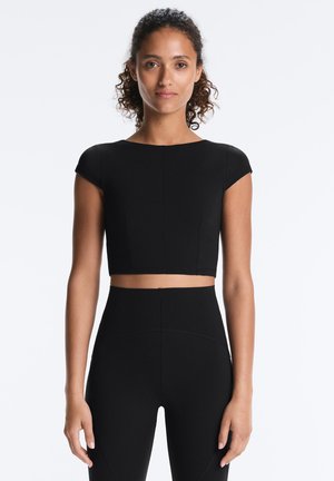 Kvinde med krøllet hår iført en sort kortærmet crop top og højtaljede sorte leggings, stående mod en ensfarvet hvid baggrund.