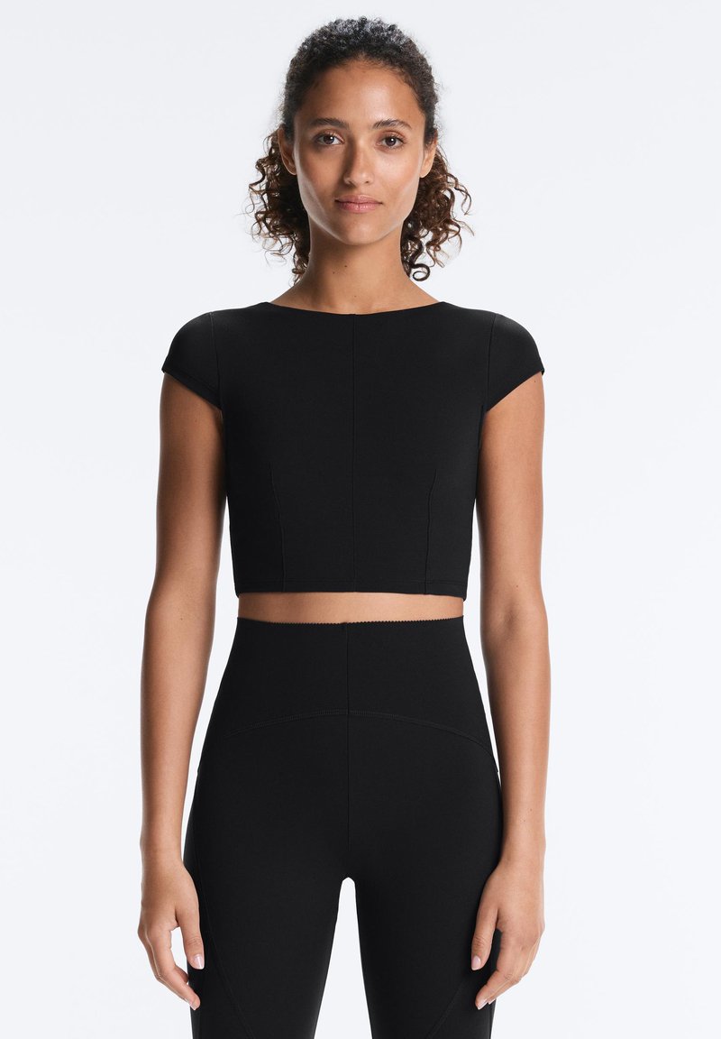 Kvinde med krøllet hår iført en sort kortærmet crop top og højtaljede sorte leggings, stående mod en ensfarvet hvid baggrund.