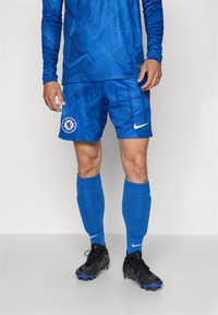 Echipament de fotbal albastru, format dintr-un tricou cu mânecă lungă, cu modele geometrice, pantaloni scurți asortați, protecții pentru tibii albastre și ghete negre cu accente albastre.