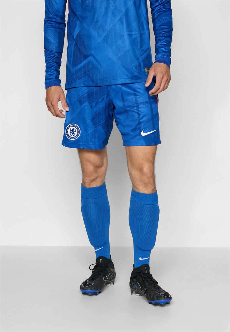 Echipament de fotbal albastru, format dintr-un tricou cu mânecă lungă, cu modele geometrice, pantaloni scurți asortați, protecții pentru tibii albastre și ghete negre cu accente albastre.