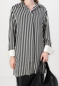 Camisa a rayas blancas y negras con cuello, mangas largas y puños blancos en contraste. La tela es suave con un acabado ligeramente brillante.