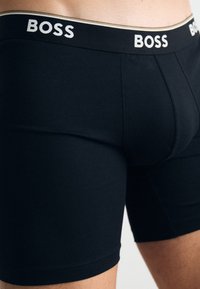 Schwarze Boxer-Briefs mit einer figurschmeichelnden Passform, ausgestattet mit einem markenbezogenen Bund in Schwarz und Gold, gefertigt aus einem glatten, dehnbaren Material.