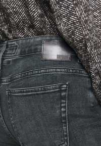 Jeans gris oscuro con una textura suave, que presentan una etiqueta de cuero que dice "DRYKORN" y costuras detalladas en los bolsillos traseros.