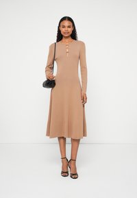 Kötött bézs hosszú ujjú ruha, fodros szegéllyel, kerek nyakkivágással és arany gombos díszítéssel. Fekete táskával és szandálokkal párosítva.