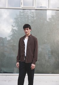 Brūna pogu krekls ar salocītu apkakli, uzvilkts virs balta T-krekla, kombinēts ar melnām biksēm uz spoguļa virsmas.