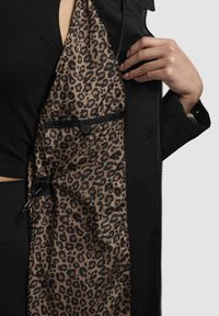 Zwarte buitenjas met een leopardprint voering. Voorzien van een ritsluiting, verstelbare trekkoord en een binnenzak met knoop.