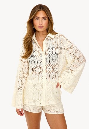 Chemisier en ivoire avec un motif hexagonal, manches longues en forme de cloche et devant boutonné, associé à un short assorti. Tissu texturé et léger.