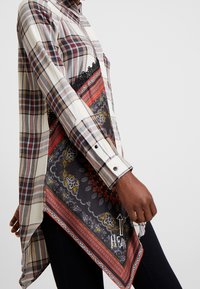 Camisa de cuadros con mangas largas, en colores crema, rojo y azul marino; presenta un borde de encaje negro y un dobladillo con un patrón similar a una bufanda que incluye detalles florales.