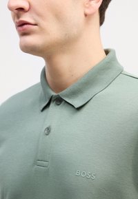 Grønn polo skjorte laget av glatt stoff, med krage, tre knapper, og en subtil "BOSS" logo på venstre bryst.