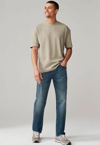 Camisa de manga corta gris claro, vaqueros de mezclilla azules, zapatillas grises con suelas y acentos blancos. Postura de pie, manos en los bolsillos.