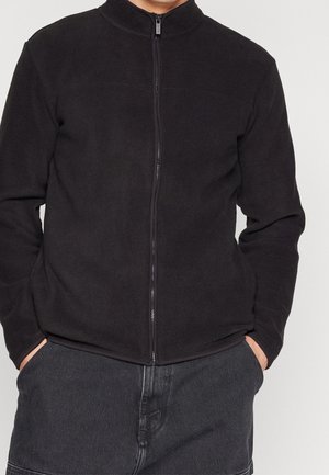 Person trägt eine schwarze Fleecejacke mit Reißverschluss und Händen in den Taschen, kombiniert mit dunkelgrauen Hosen vor einfarbigem Hintergrund.