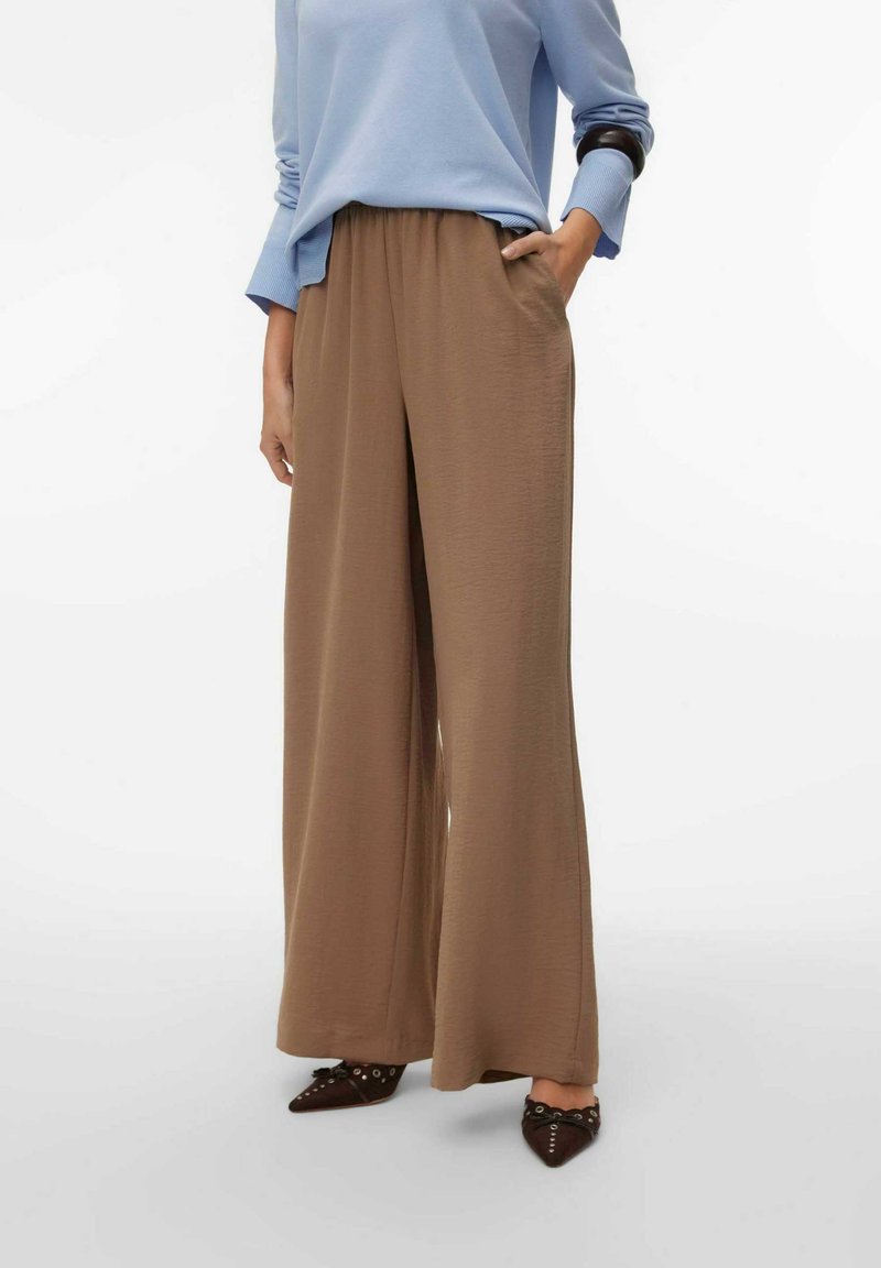 Pantalon marron à jambes larges avec une taille élastique et une texture lisse. Associé à un haut à manches longues bleu clair et des chaussures pointues foncées.