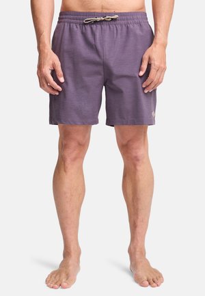 Man draagt paarse short met trekkoord, staat blootsvoets tegen een effen achtergrond, handen ontspannen langs de zij.