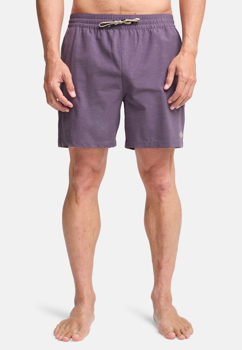 Άνδρας που φοράει μωβ σορτς με κορδόνι, στέκεται ξυπόλητος μπροστά σε απλό φόντο, με τα χέρια χαλαρά στο πλάι.