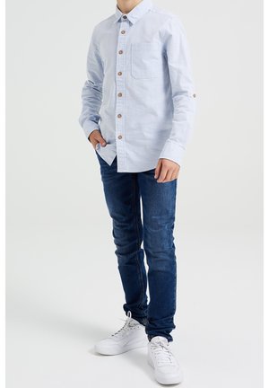 Helles blaues Langarmhemd mit Knopfverschluss, Brusttasche und kontrastierenden braunen Knöpfen, kombiniert mit dunkelblauen Jeans und weißen Sneakern.