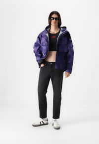 Purple camo jacket with gray lining, black cropped top, black jeans, and gray sneakers with black stripes. Model wears sunglasses.