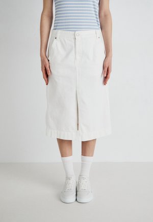 Person iført en hvid knælængde nederdel med frontslids, hvide sneakers, hvide sokker og en lyseblå striped top, stående på en ensfarvet gulv.