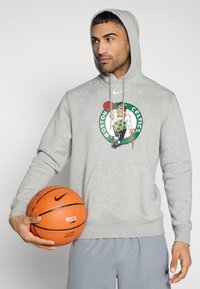 Γκρι φούτερ με το λογότυπο των Boston Celtics· διαθέτει τσέπη καγκουρό και ρυθμιζόμενο κορδόνι. Το μοντέλο κρατάει μια πορτοκαλί μπάλα μπάσκετ Nike.