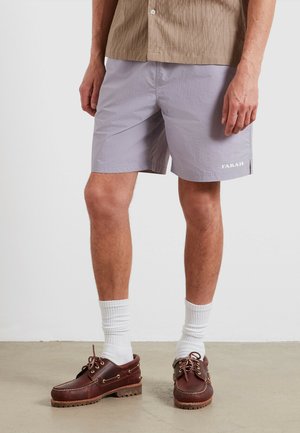Mężczyzna w jasnofioletowych szortach z logo "FARAH", białych skarpetkach do kostki, brązowych skórzanych butach i beżowej koszuli z guzikami.
