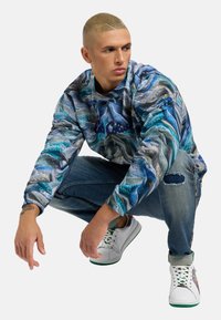 Sudadera con un estampado colorido en azul y gris, jeans azules desgastados y zapatillas blancas con acentos en verde, usadas por una persona en una pose de cuclillas.