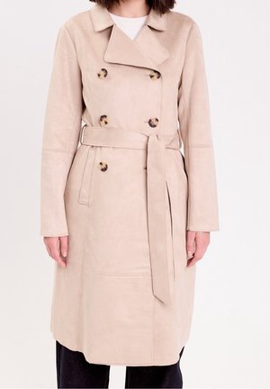 Femme portant un trench-coat beige mi-long avec ceinture et boutons en écaille, sur un haut blanc et un pantalon noir.