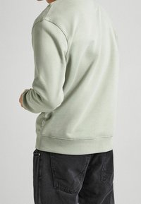 Ljusgrön sweatshirt med rund halsringning och ribbade ärmslut, tillsammans med mörkgrå byxor. Mjuk tygkvalitet och avslappnad passform.