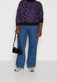 Lila und schwarze gemusterte Sweater, lockere blaue Jeans mit Seitentasche, schwarze Handtasche und graue Sneaker mit farblichen Akzenten.