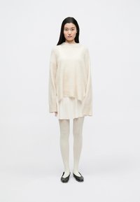 Pulover supradimensionat, bej deschis, cu manșete late, purtat peste o rochie crem. Asortat cu dresuri deschise și balerini negri. Textură simplă, moale.