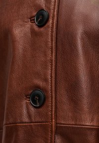 Κοντινό πλάνο καφέ δερμάτινου σακακιού, με ανάγλυφο υλικό, έντονη ραφή και δύο μαύρα κουμπιά εμπρός για κλείσιμο.