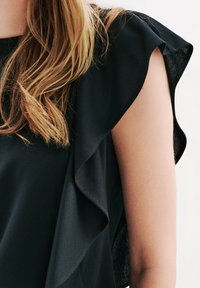 Svart topp med volangdetaljer på ärmarna, tillverkad av ett slätt tyg. Urringningen är rundad och texturen verkar mjuk mot huden.