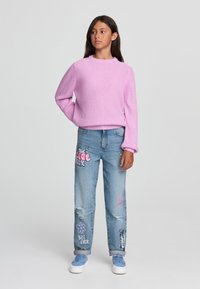 Maglione rosa lavorato a maglia con scollatura a girocollo, abbinato a jeans blu strappati con stampe grafiche colorate e orli arrotolati. Sneakers bianche.