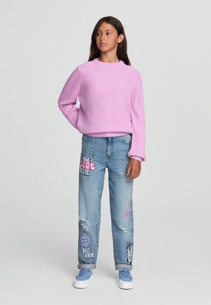 Maglione rosa lavorato a maglia con scollatura a girocollo, abbinato a jeans blu strappati con stampe grafiche colorate e orli arrotolati. Sneakers bianche.