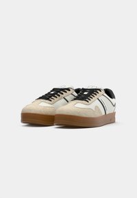 White and beige sneakers with black laces, suede and leather materials, featuring a gum rubber sole and branded side detail.