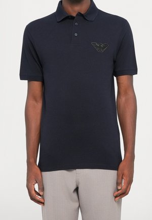 Mann trägt ein schwarzes kurzärmeliges Poloshirt mit einem kleinen gestickten Logo auf der linken Brust und hellgraue Hose.