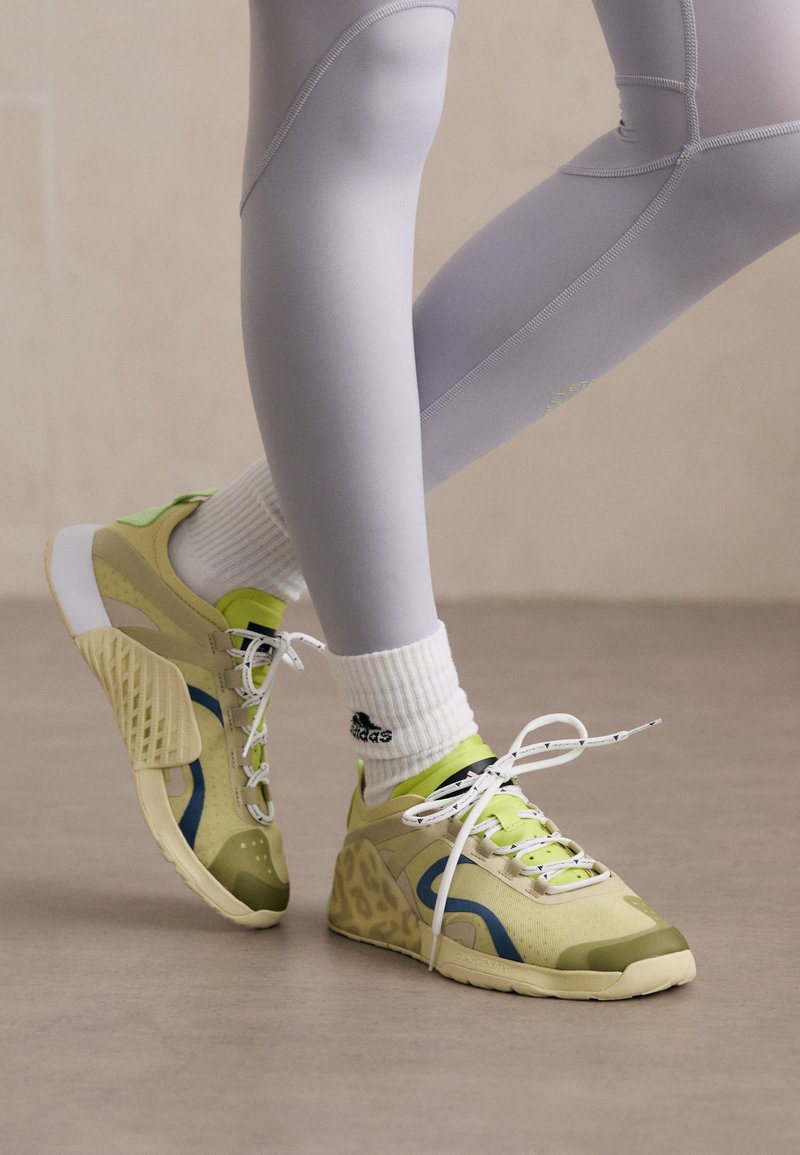 Gaiši zaļi un bēši sporta apavi ar ziliem akcentiem un teksturētām daļām, ar baltām auklām un mīkstinātu papēdi atbalstam.