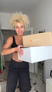 Een persoon houdt twee pakketten vast, één bruin met een oranje logo en één witte. De setting is een kamer met lichtgekleurde muren en een spiegel.