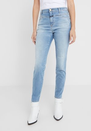 Woman wearing light blue slim-fit jeans with subtle distressing and white ankle boots, standing against a plain light background.