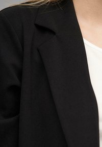 Fekete blézer sima textúrával és letisztult dizájnnal, fehér, bordázott felsővel párosítva. Kiemelten a hajtóka és a váll részleteire.
